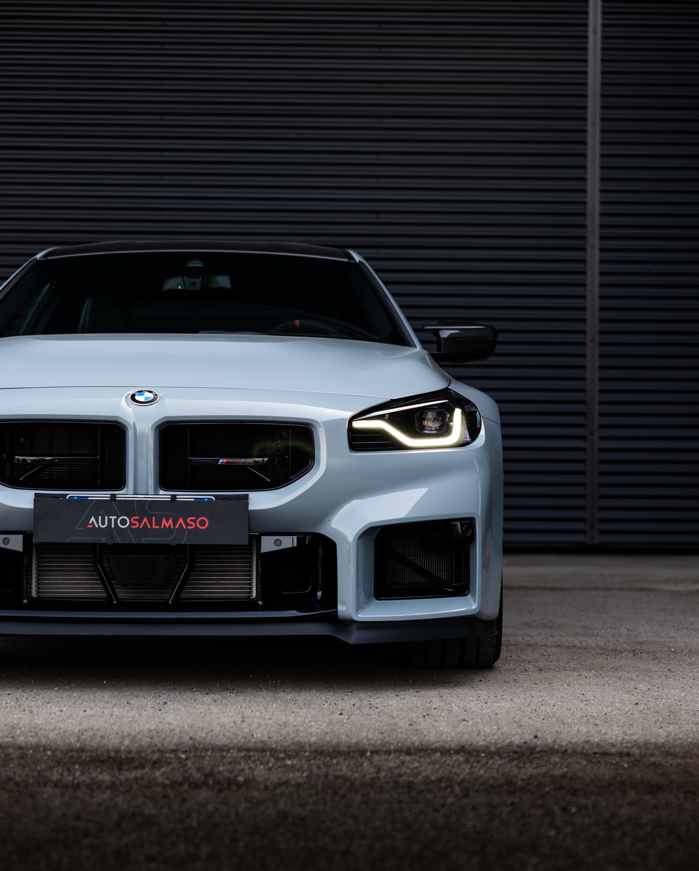 Frontale di BMW M2 grigio chiaro con fari LED accesi e portatarga AUTOSALMASO davanti a una saracinesca scura